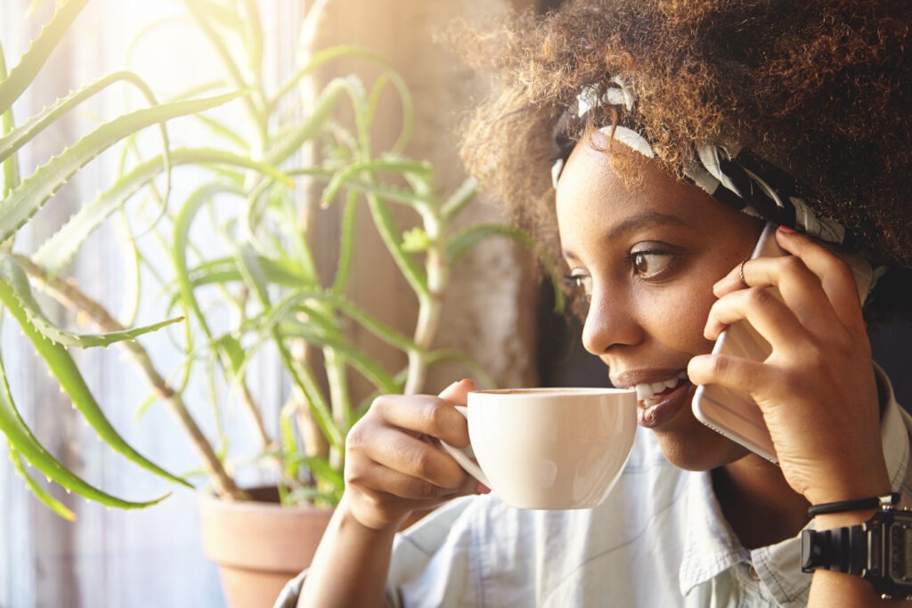 Portrait of thoughtful African woman with Afro haircut, wearing trendy clothes resting at home, looking through window, holding cup of coffee while talking on cell phone, inviting friends to her place